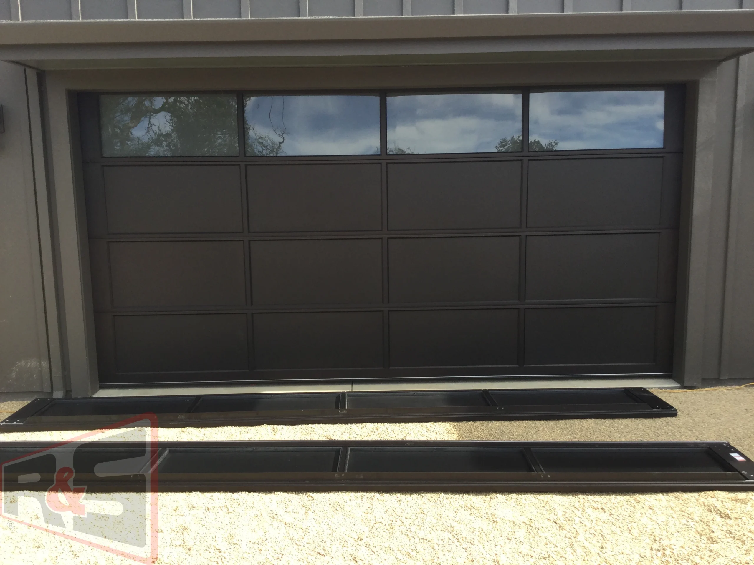 A dark modern sectional garage door with top glass panels and detached replacement sections on the ground addressing Garage Door Problems Vallejo.